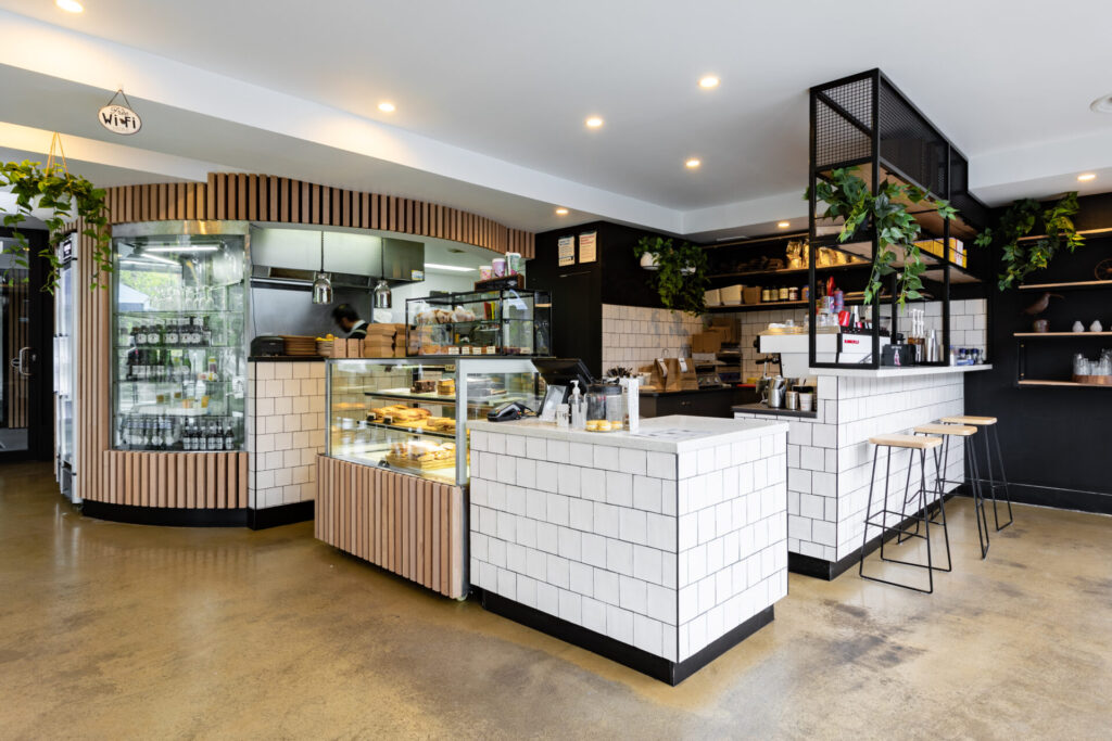 Modern Kafe interior near Denmark Hill with white tiled counters, wooden accents, and indoor plants. Glass display cabinets show pastries and drinks. Shelves with mugs line the black wall, and two bar stools sit at the counter. Hillbrook Projects