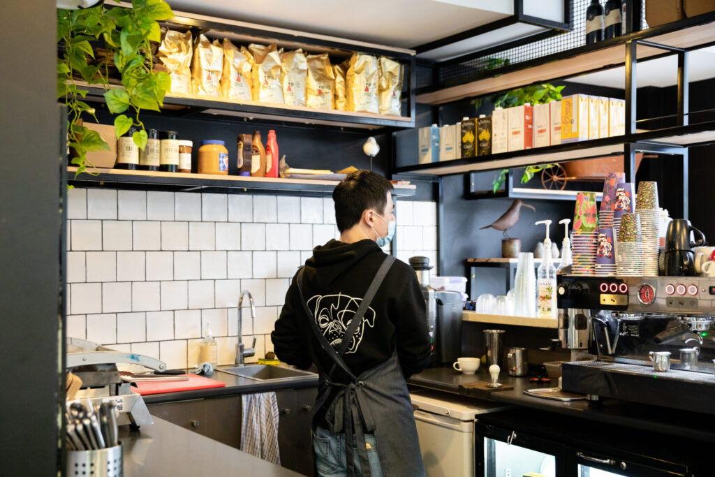 A barista wearing a black hoodie and apron stands behind the coffee bar at Denmark Hill Kafe, surrounded by coffee machines, cups, and shelves stocked with coffee bags and supplies in a modern café. Hillbrook Projects