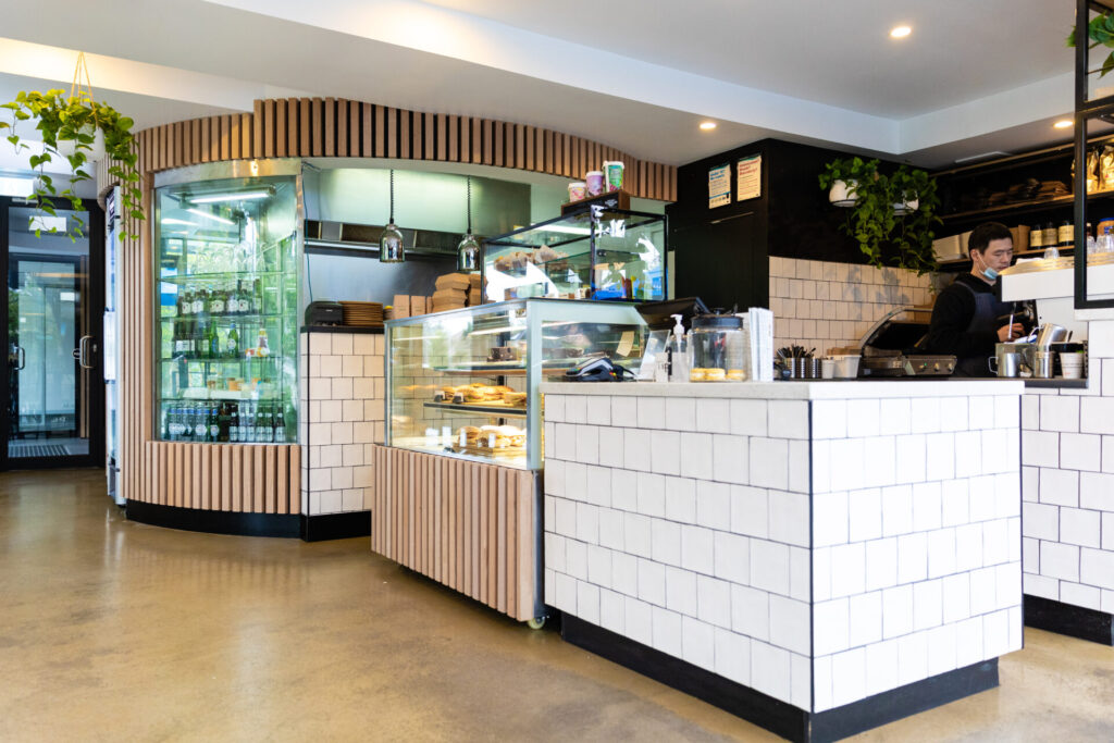 Modern Kafe interior near Denmark Hill, featuring white tiled counters, glass display cases filled with pastries, drinks in the fridge, a barista preparing coffee, and lush green plants hanging from the ceiling. The space is bright and clean. Hillbrook Projects