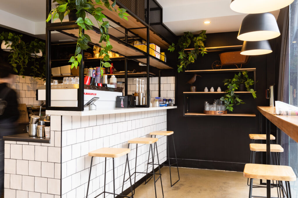 A modern café interior near Denmark Hill, featuring white tiled counters, wooden stools, hanging plants, open shelves with cups and decor, and large windows letting in natural light. Hillbrook Projects