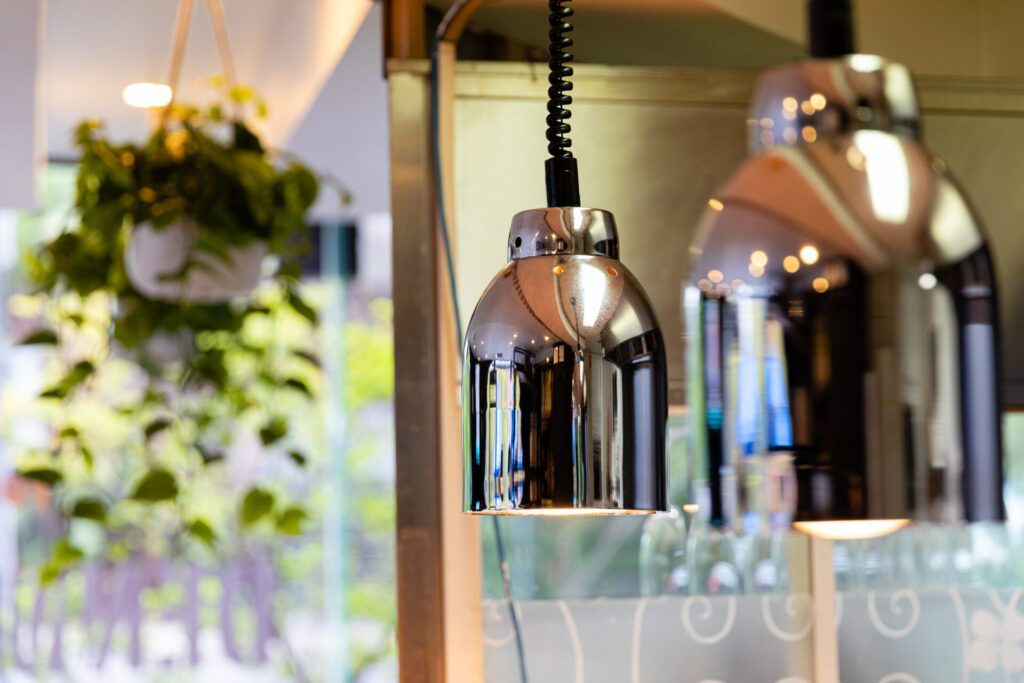 Close-up of shiny, metallic hanging pendant lamps at Kafe, Denmark Hill, with a blurred background featuring green hanging plants and soft natural light. Hillbrook Projects