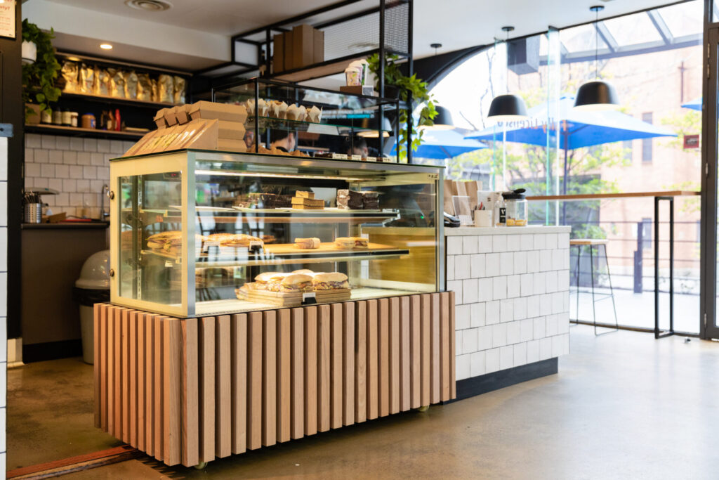 A modern Denmark Hill café interior with a wooden display counter filled with pastries and sandwiches, white tiled surfaces, hanging lights, and large windows letting in natural light. Blue umbrellas are visible outside. Hillbrook Projects
