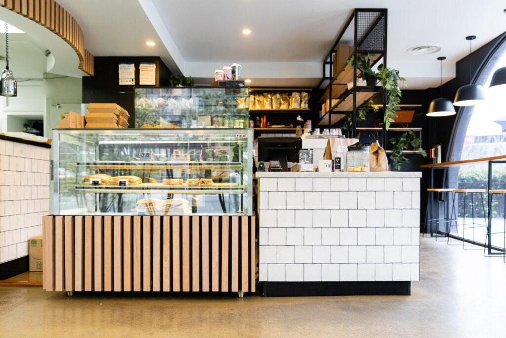 Modern Kafe interior near Denmark Hill with a glass display of pastries, white tiled counter, shelves filled with snacks and plants, and large windows letting in natural light. The space appears clean and welcoming. Hillbrook Projects