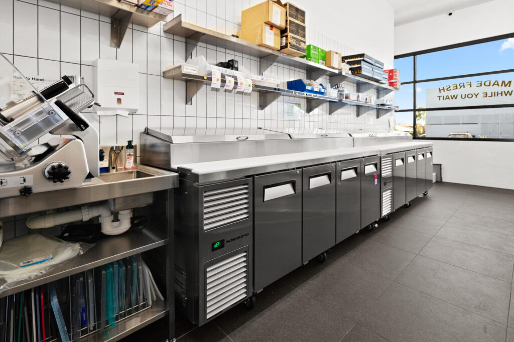 A clean, modern commercial kitchen with stainless steel worktops and prep equipment—ideal for creating unforgettable Grazing Moments. Shelves above hold various boxes and supplies, while large windows fill the space with natural light. Hillbrook Projects