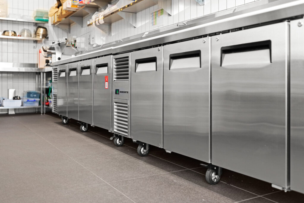 A row of stainless steel Grazing Moments commercial fridges with wheels lines the wall of a clean, modern kitchen. Shelving above holds kitchen supplies and utensils, while the floor is finished in dark tiles. Hillbrook Projects