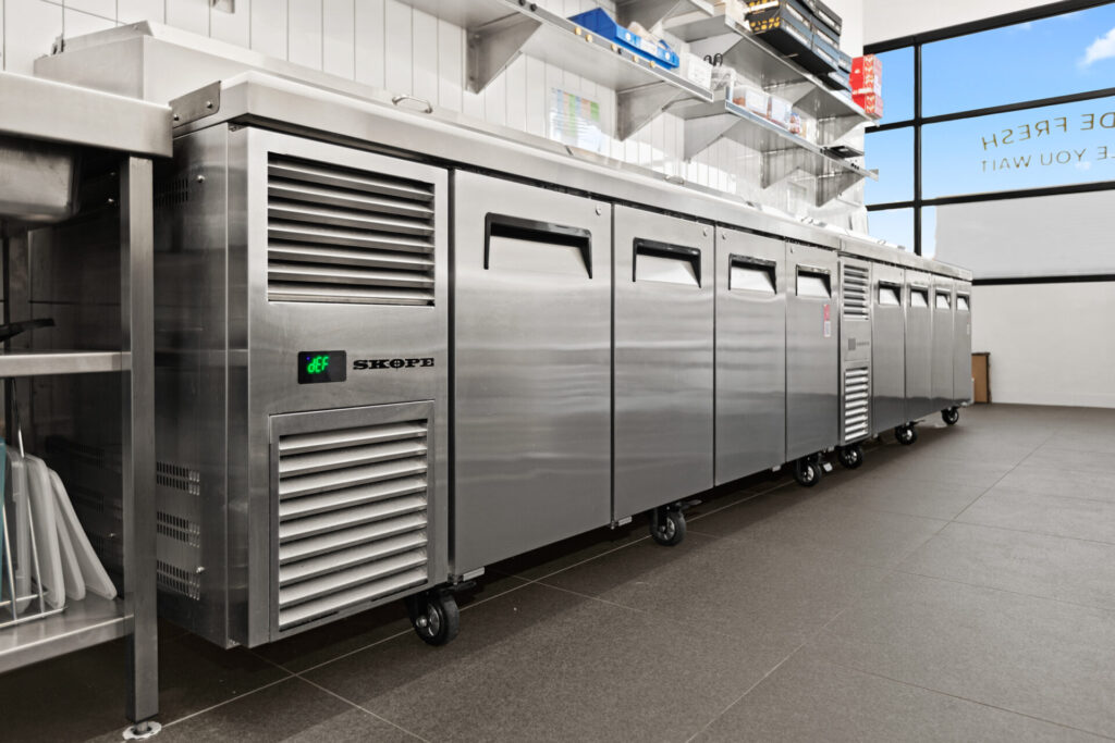 A row of commercial stainless steel fridges and freezers on wheels lines a clean, modern kitchen from Grazing Moments, with tiled walls, shelves, and large windows providing natural light. Hillbrook Projects