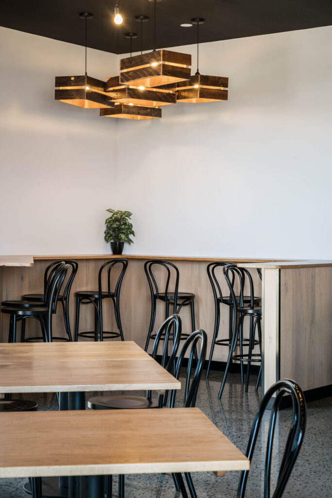 Modern café interior with wooden tables, black chairs, and a geometric pendant light fitting. A small potted plant sits on the counter of Portz Kebab & Grill, creating a minimalist and inviting atmosphere against a plain white wall. Hillbrook Projects