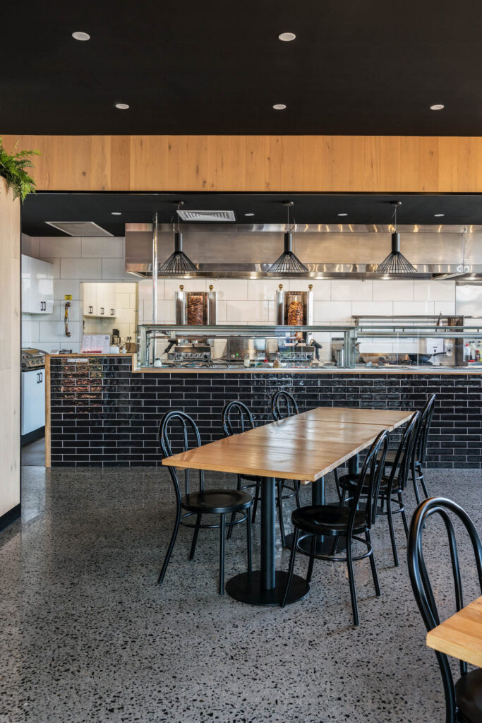Modern restaurant interior with wooden tables, black metal chairs, and a sleek black tile counter. The open kitchen at Portz Kebab features stainless steel appliances, a visible grill, hanging pendant lights, and a polished concrete floor. Hillbrook Projects