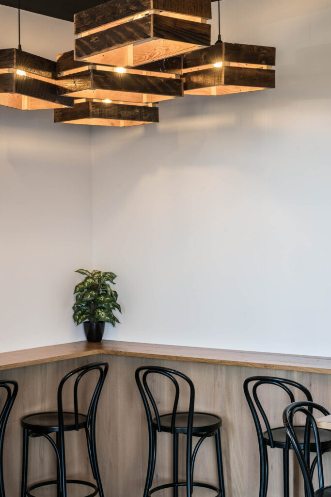 A minimalist café corner with wooden bar counters, black metal stools, a small potted plant, and geometric wooden pendant lights creates a cosy spot to enjoy a fresh Portz Kebab hot off the grill against a crisp white wall. Hillbrook Projects