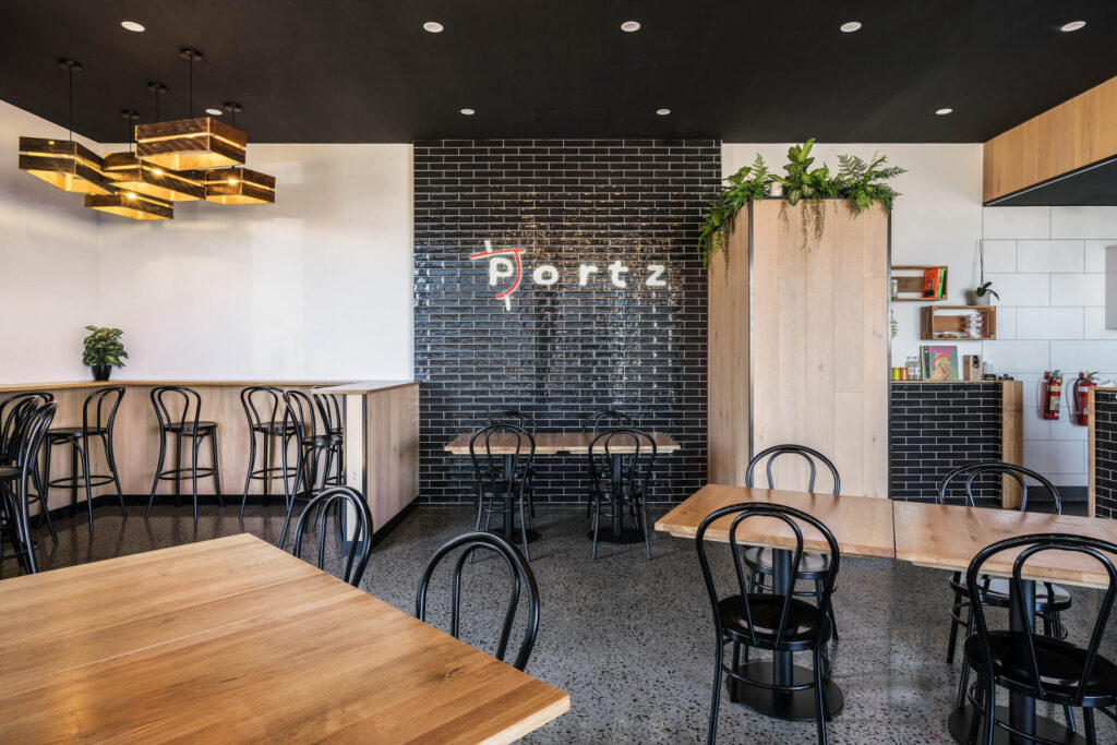 A modern café interior with wooden tables, black chairs, a black brick feature wall displaying "Portz Kebab & Grill," and geometric pendant lights. Green plants add a touch of colour to the stylish, minimalist space. Hillbrook Projects