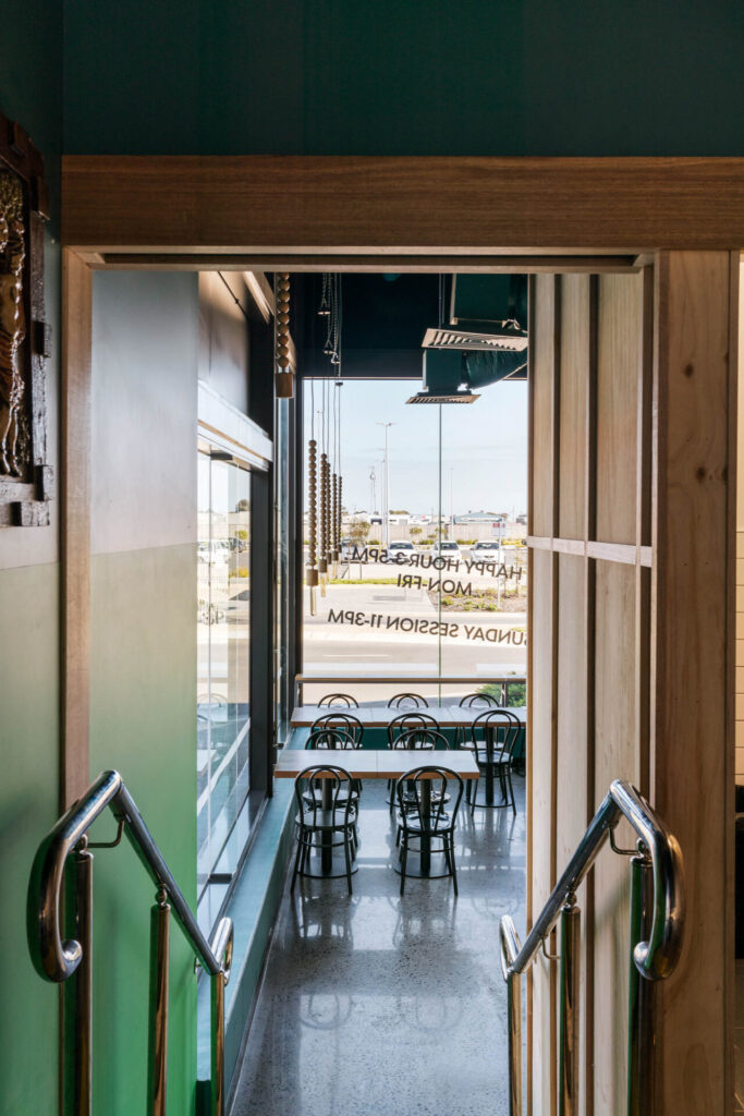 View through a doorway to the cosy Little Raaja cafe seating area, with several tables and black chairs by large windows streaming in natural light; an outdoor car park and street visible beyond. Hillbrook Projects