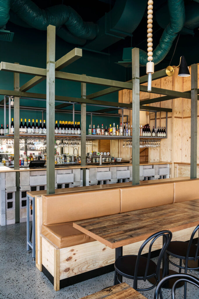 A modern bar with green accents, wooden shelves, and a variety of bottles behind the counter. Tan cushioned bench seating and rustic wooden tables create a cosy vibe at Little Raaja, complemented by black chairs and industrial décor elements. Hillbrook Projects