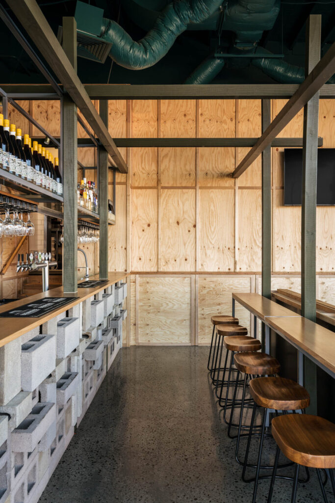 Little Raaja is a modern bar with a concrete and plywood interior, featuring a long wooden bar with stools, shelves of bottles and glasses, and exposed green ventilation ducts on the ceiling. Hillbrook Projects