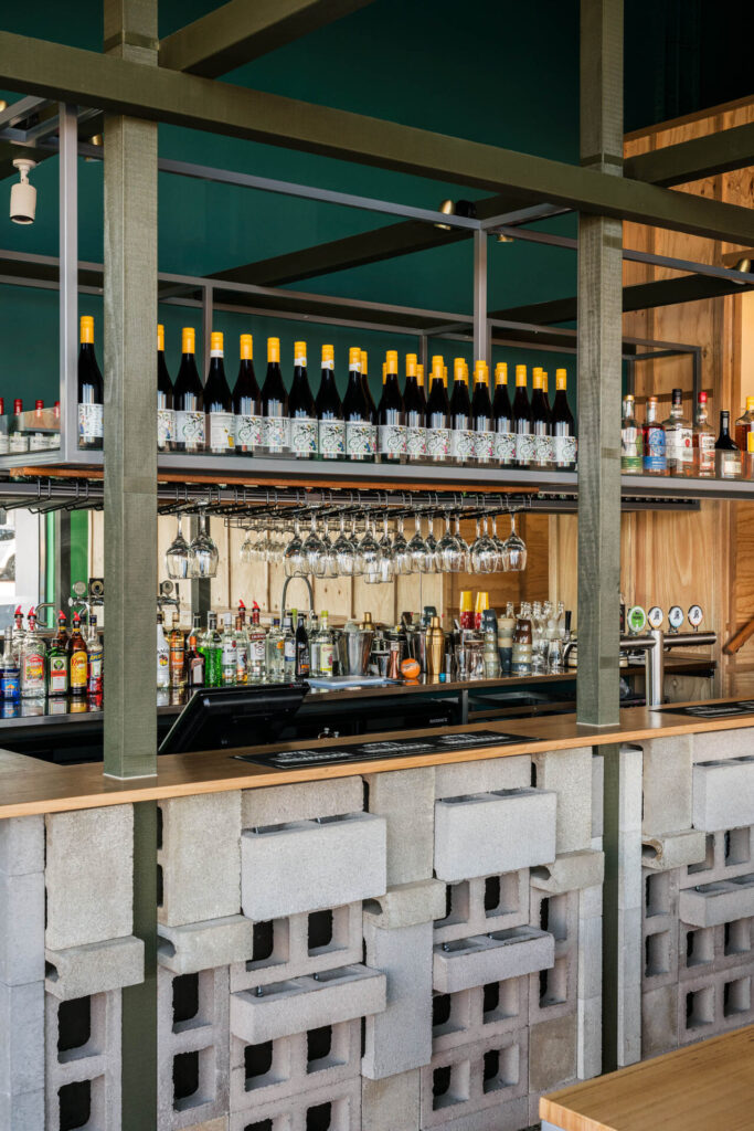 A modern bar, Little Raaja, features a counter made of stacked concrete breeze blocks, shelves lined with wine bottles, hanging wine glasses, and a backdrop of spirits bottles and drink taps. Hillbrook Projects