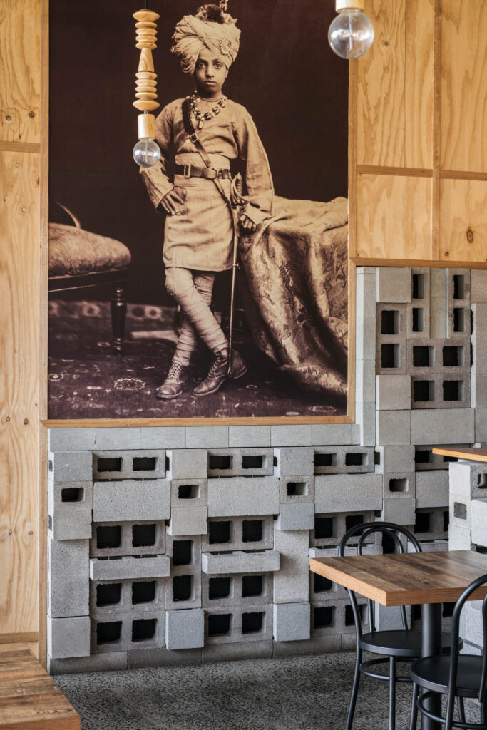 A sepia-toned portrait of Little Raaja in traditional attire hangs above a modern café with unfinished breeze block walls, wooden panels, black chairs, and wooden tables. Suspended light fittings add a contemporary touch. Hillbrook Projects