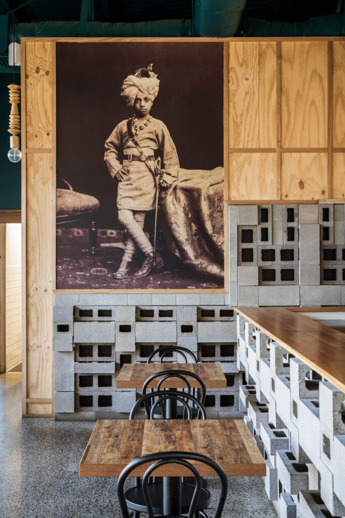 A café interior with wooden tables, black chairs, breeze block decor, and a large sepia-toned vintage portrait of Little Raaja—a child in fancy dress—on the wall. Hillbrook Projects