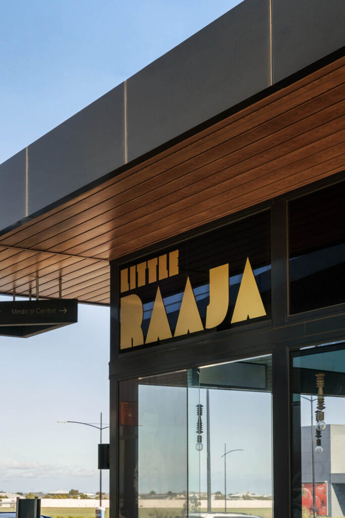 A stylish shopfront features a black and gold “LITTLE RAAJA” sign beneath a modern wooden awning, with reflections and street lamps dancing in the glass windows, inviting you into the world of Little Raaja. Hillbrook Projects