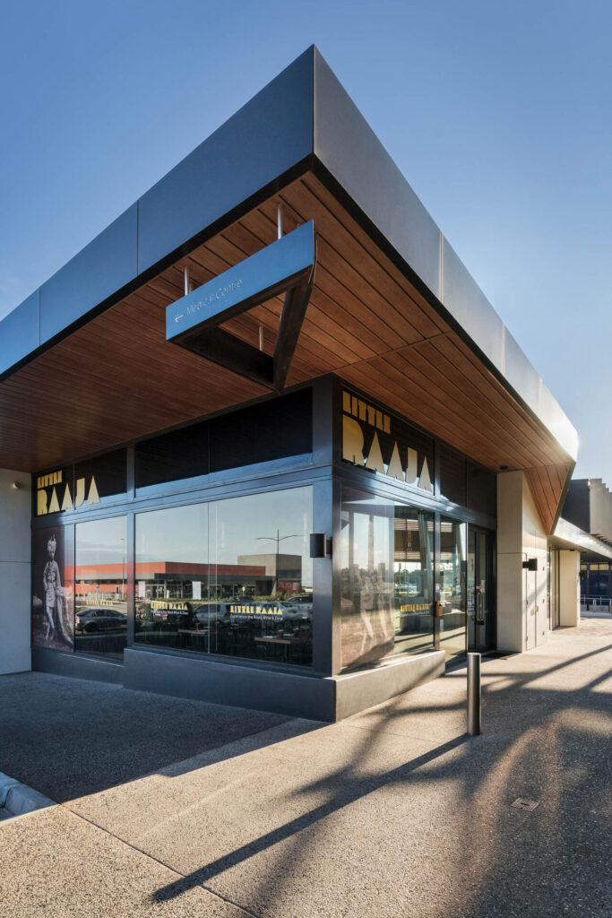 Modern commercial building with large glass windows, wooden ceiling accents, and bold signage reading "LITTLE BAAJA." The rectangular structure beneath a clear blue sky hints at the vibrant charm of Little Raaja. Hillbrook Projects