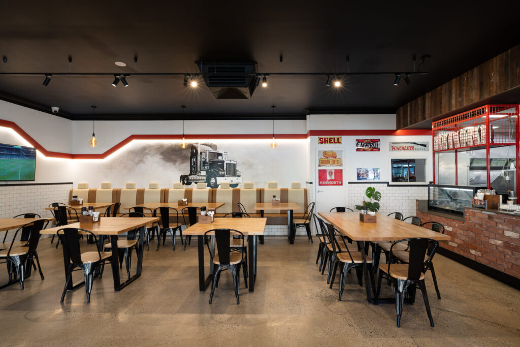 Modern restaurant interior at Corio Bay Roadhouse with wooden tables, black metal chairs, and a counter with red accents. A mural of a lorry decorates the back wall, whilst posters and shelves display various items. Space appears bright and clean. Hillbrook Projects