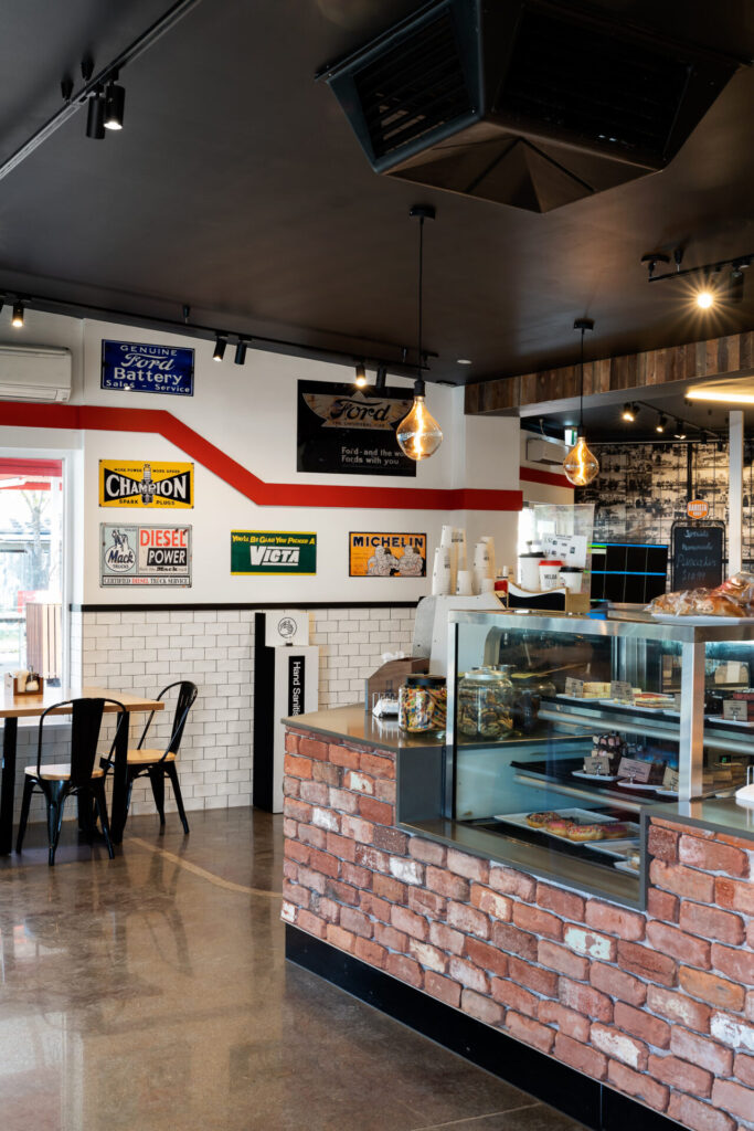 A modern café interior with a glass counter displaying pastries, vintage Roadhouse car signs on white walls, hanging light bulbs, and a table with black chairs by a window overlooking Corio Bay. Hillbrook Projects