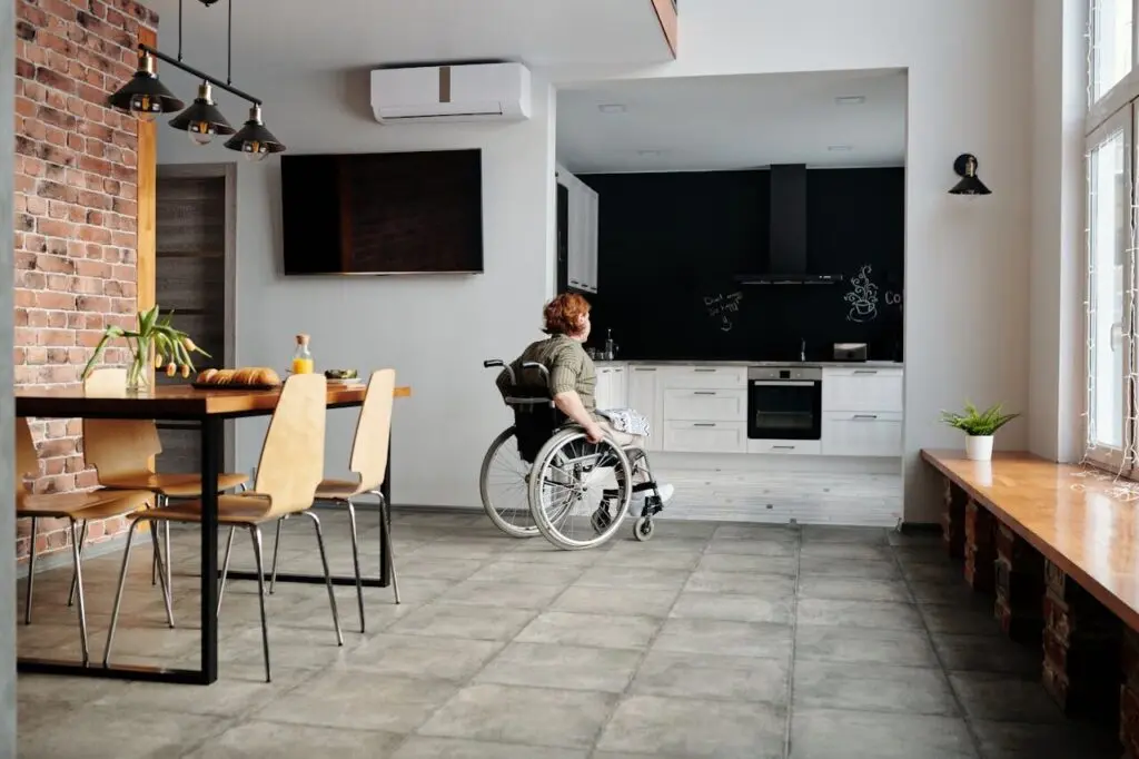A person in a wheelchair moves towards a modern kitchen in an open, spacious Specialist Disability Accommodation flat with brick walls, large windows, a dining table, and plenty of natural light. Hillbrook Builder