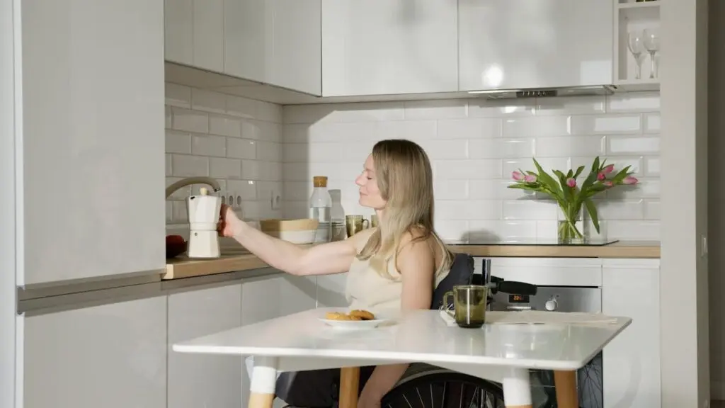 A woman in a wheelchair sits at a kitchen table within Specialist Disability Accommodation, holding a moka pot. The modern kitchen features white cupboards, pink tulips, and an array of food and drinks on the table. Hillbrook Builder