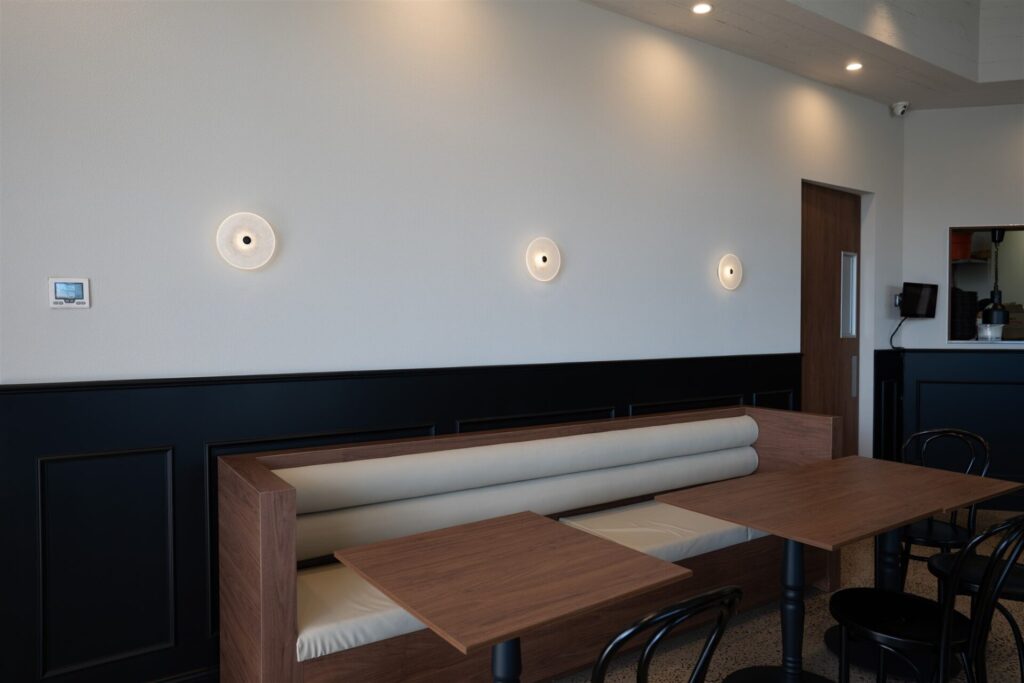 A modern café interior at Town & Country Waurn Ponds features wooden tables, a cushioned bench, black chairs, and three round wall lights above. The white wall with black panelling enhances the space’s minimalist design. Hillbrook Projects