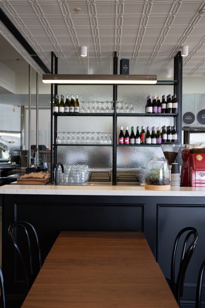 A view of the modern Town & Country Waurn Ponds café bar with glass shelves displaying wine bottles and glassware, a countertop with glasses, and a wooden table with black chairs in the foreground. Hillbrook Projects