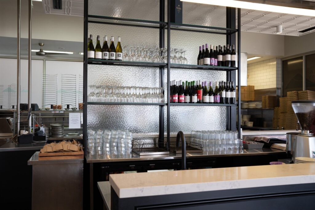 A bar area in a modern Waurn Ponds restaurant features shelves of wine bottles and glasses. In the background, a kitchen with pizza boxes and metal racks is visible, while a coffee machine sits front and centre on the counter at Town & Country. Hillbrook Projects
