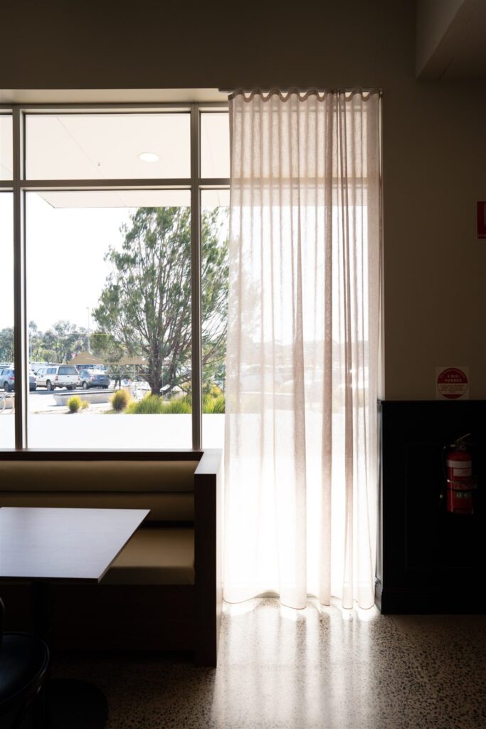 A softly lit café corner with a wooden booth and table sits beside a large window at Town & Country Waurn Ponds, sheer white curtains letting in natural light and offering a view of trees and parked cars outside. Hillbrook Projects