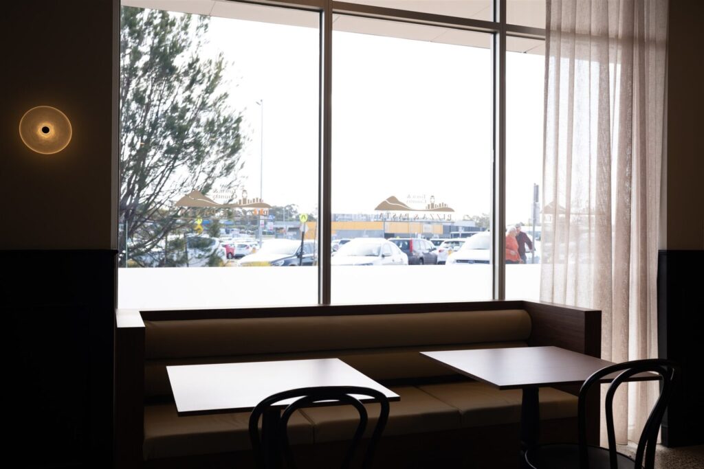 A modern café at Town & Country Waurn Ponds features empty tables and chairs near large windows. Sheer curtains let in daylight, several cars are parked outside, and a small round wall light glows inside. Hillbrook Projects