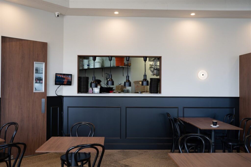 A modern café in Waurn Ponds with black chairs and wooden tables, featuring a service hatch into the kitchen, black panelling on white walls, and a single cup on a table. Warm ceiling lights glow above, whilst a security camera keeps watch. Hillbrook Projects