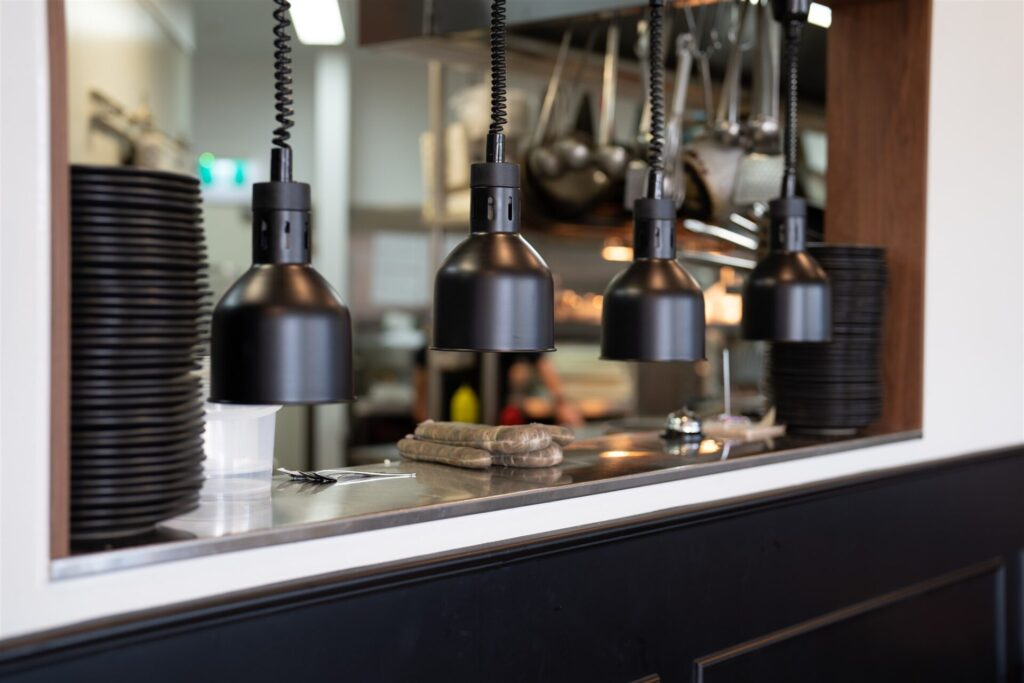 A restaurant kitchen pass at Town & Country Waurn Ponds features four black heat lamps above a counter lined with stacked black plates and a cloth, with the bustling kitchen and utensils visible in the background. Hillbrook Projects