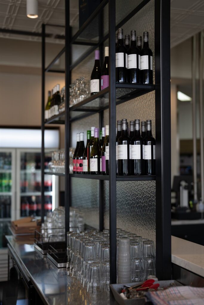 Bottles of wine, drinking glasses, and stacked glassware are neatly arranged on black metal shelves in the modern Town & Country Waurn Ponds bar, with a fridge in the background. Hillbrook Projects