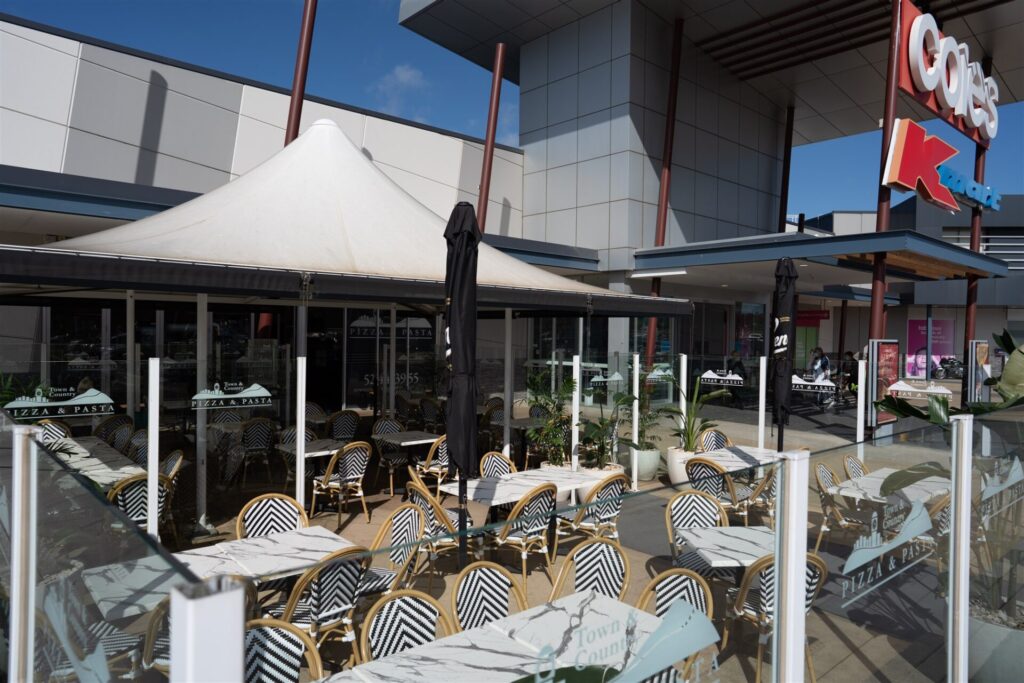 Outdoor seating area of a café with black-and-white striped chairs and several tables, enclosed by glass panels. A large white canopy provides shade, with a Coles supermarket sign and Town & Country Waurn Ponds visible in the background. Hillbrook Projects