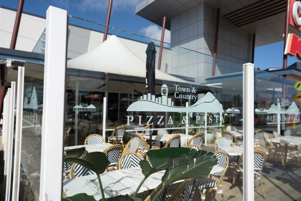 Relax in the outdoor seating area at Town & Country Waurn Ponds, featuring white tables and chairs beneath a white canopy. A glass panel displays the logo, set against a backdrop of a modern building. Hillbrook Projects