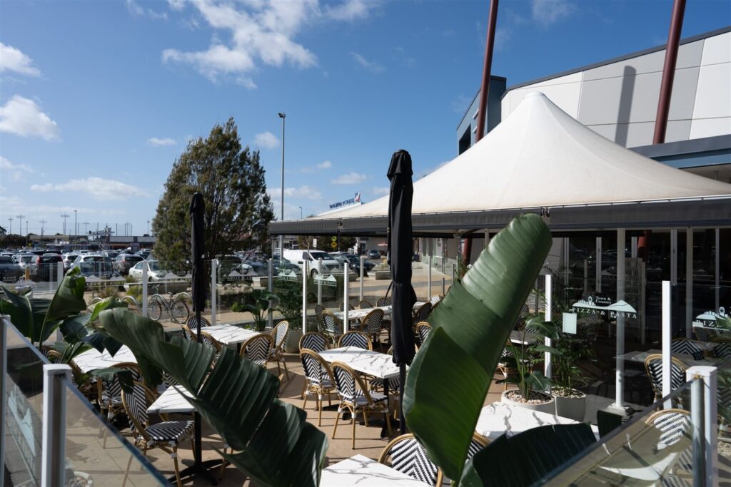 Outdoor café patio with black and white chairs and tables, surrounded by green plants and glass barriers under a white canopy at Town & Country, Waurn Ponds. Car park and buildings are visible in the background on a sunny day. Hillbrook Projects