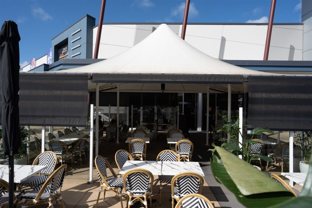 Outdoor café at Town & Country, Waurn Ponds, with black-and-white chevron chairs and white tables under a large white canopy. Some tables are shaded by the canopy, surrounded by plants. Modern building and clear blue sky complete the scene. Hillbrook Projects