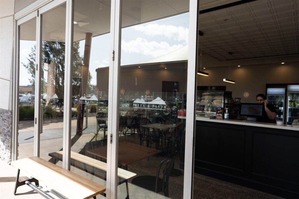 Glass doors of Town & Country Waurn Ponds restaurant with "Pizza & Pasta" signage reflect trees and parked cars outside. Inside, a staff member stands behind the counter, with empty tables and chairs visible. Hillbrook Projects