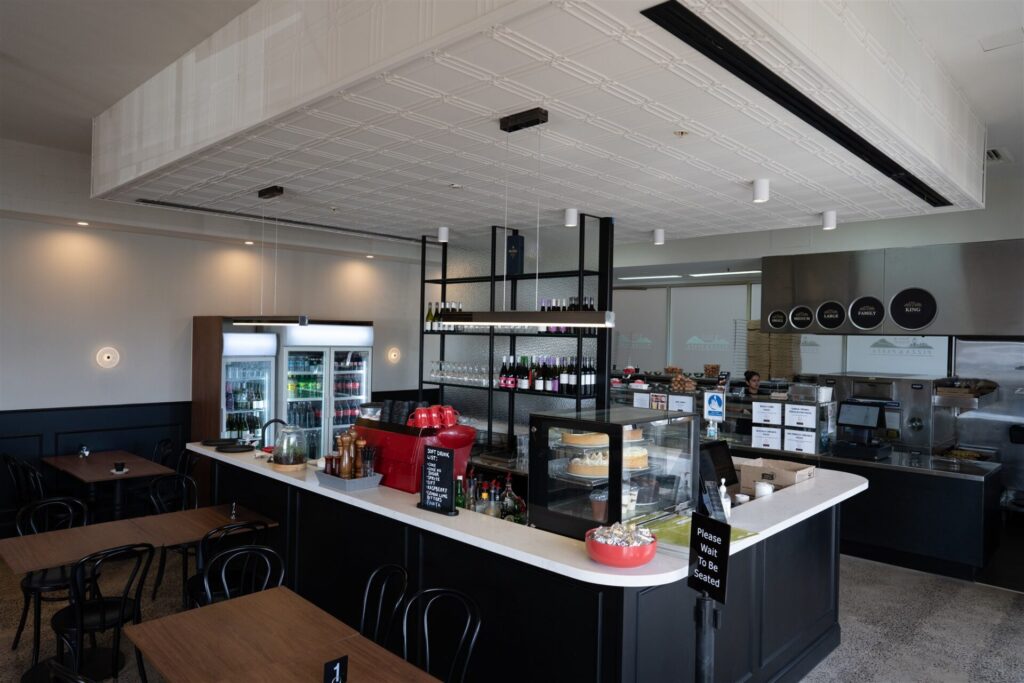 A modern café interior at Town & Country Waurn Ponds features black and white décor, display fridges with drinks, a counter with cakes and coffee machines, plus neatly arranged tables in a clean, bright space with contemporary design. Hillbrook Projects