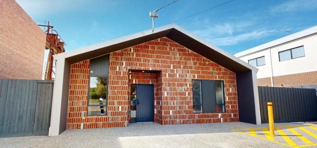 A modern brick house with a gabled roof, large black-trimmed windows, and a central dark door. The building is surrounded by a fenced garden, a yellow bollard, and a blue sky overhead. Hillbrook Projects