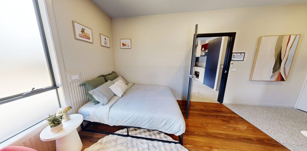 A cosy bedroom with a neatly made bed, green and white pillows, artwork on the walls, a bedside table with a plant, wooden floors, and an open door leading to a hallway and a kitchen area. Hillbrook Projects