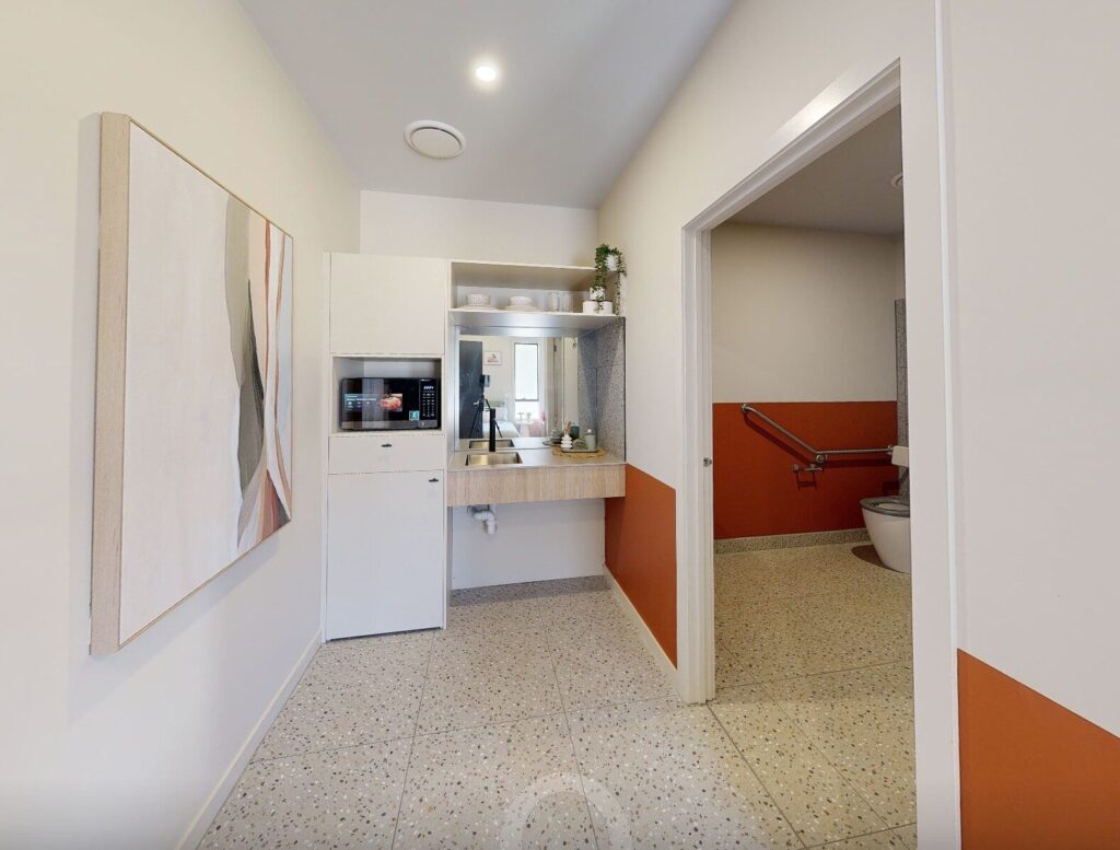 A small kitchenette with a sink, microwave, and mini-fridge next to a modern art painting; an accessible bathroom with safety rails is visible through an open doorway. The decor includes white and orange walls with speckled flooring. Hillbrook Projects
