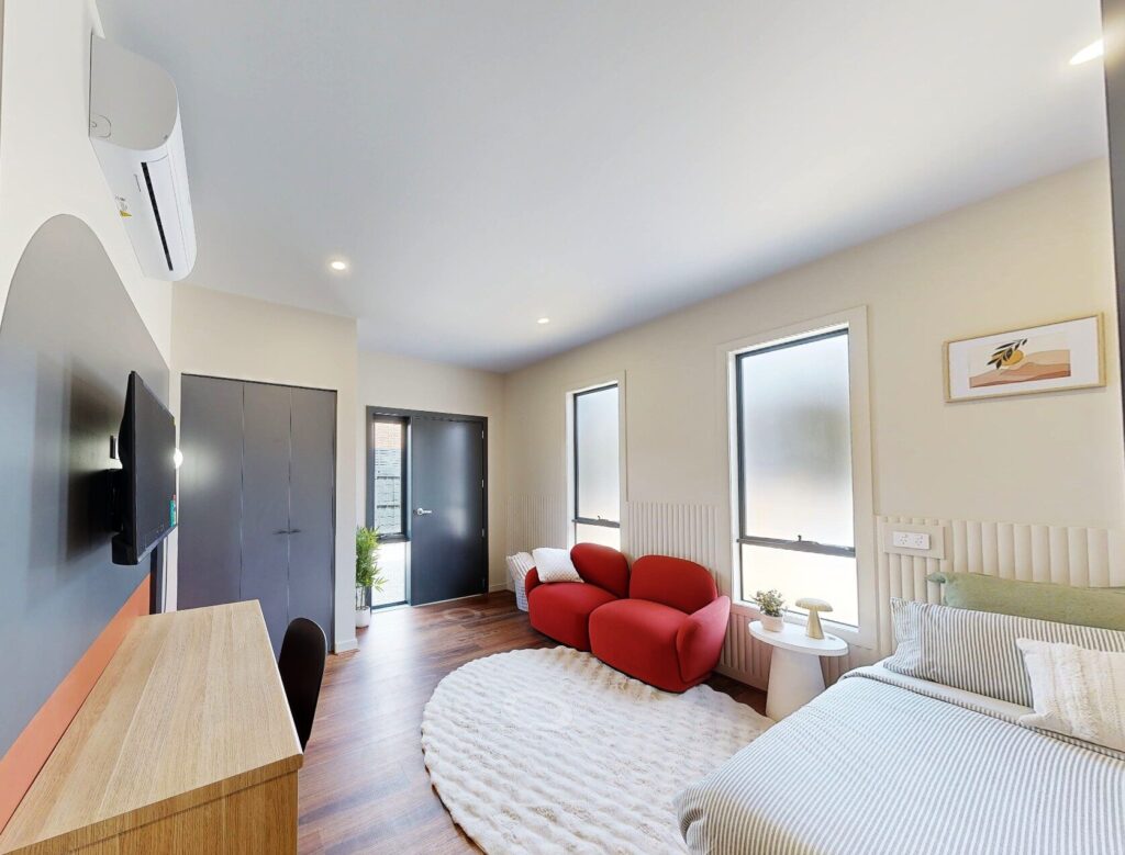 A modern, well-lit bedroom with a bed, red armchair, round white rug, wooden desk, wall-mounted TV, two windows, a plant, and a black double-door wardrobe. The decor is minimal and contemporary. Hillbrook Projects
