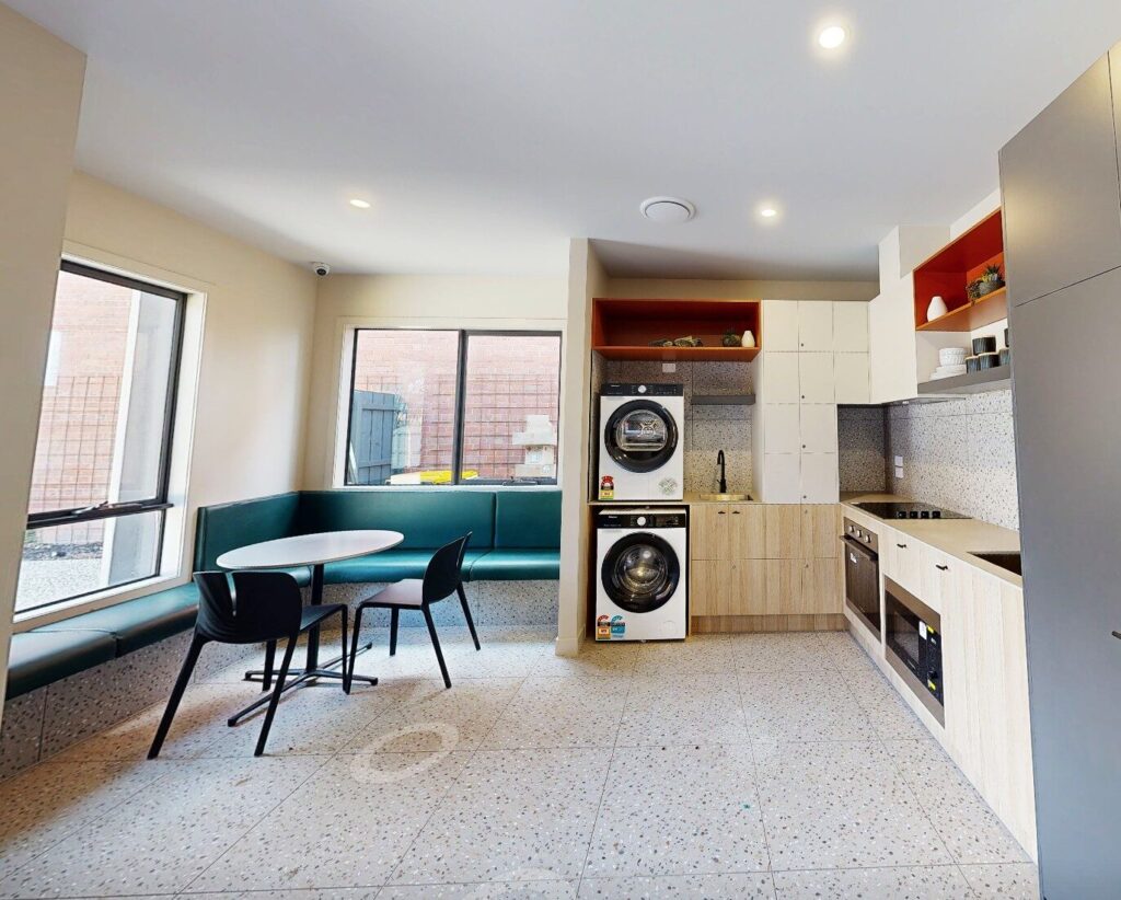 A modern kitchen with light wood cupboards, a built-in oven, stacked washer and dryer, and a round table with black chairs beside a green upholstered bench under large windows. Hillbrook Projects