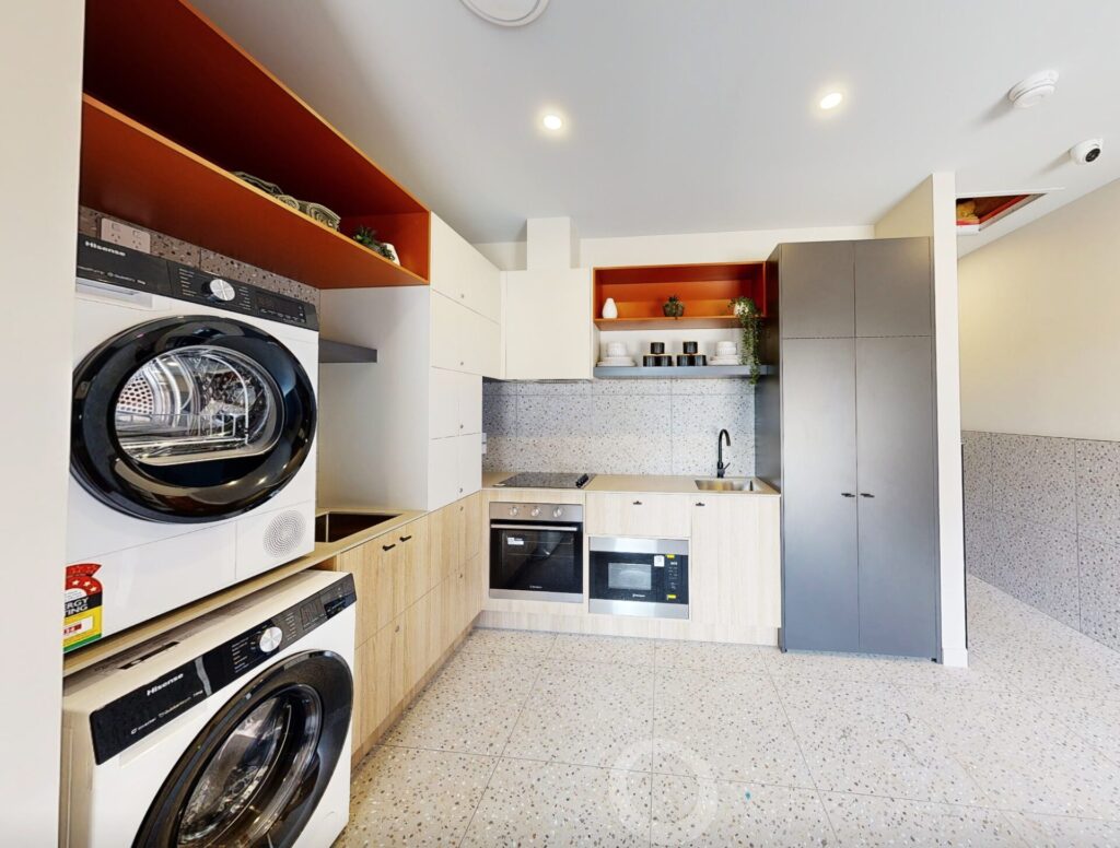 Modern laundry and kitchen area with light wood cabinets, grey worktops, a stacked washer and dryer, built-in oven, microwave, sink, and open shelves with crockery and decor. Bright lighting and speckled floors throughout. Hillbrook Projects