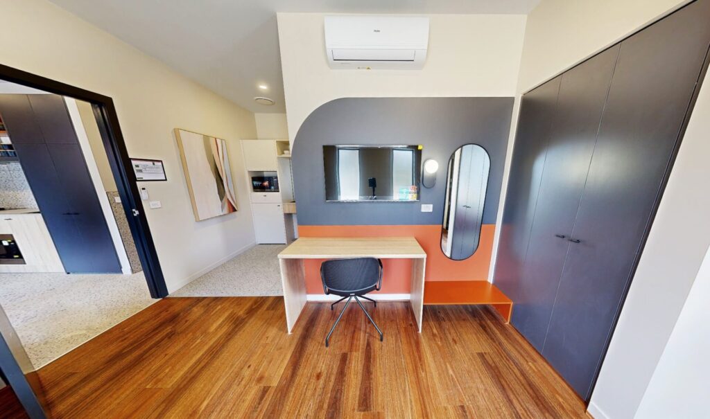 A modern room with wooden floors, a desk and chair facing a wall with a round mirror and oval mirror, built-in dark wardrobes, and an open doorway leading to a kitchen area. Hillbrook Projects